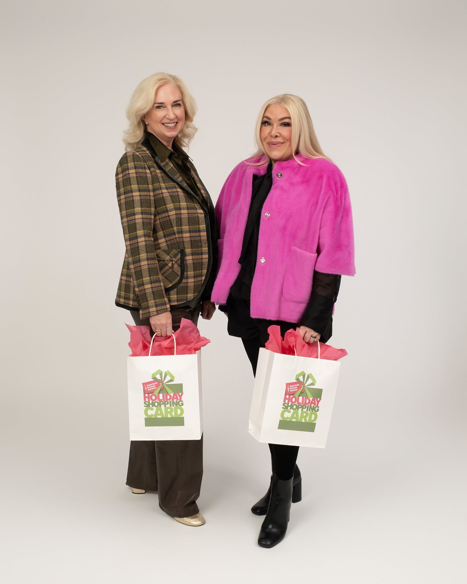 2026 Holiday Shopping Card Co-Chairs Suzanne Stiles and Lara Bell, smilling and holding Holiday Shopping Card shopping bags, which are filled with pink tissue paper. 